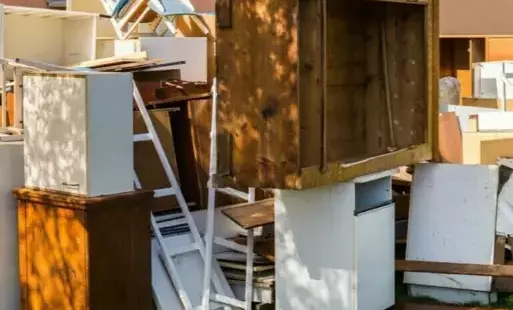 Obraz przedstawia stos starych, zniszczonych mebli i innych przedmiotów zgromadzonych na zewnątrz. Widoczne są szafki, drewniane elementy, schody i inne fragmenty wyposażenia. Meble są w różnym stanie, niektóre są uszkodzone, inne brudne i zniszczone prze