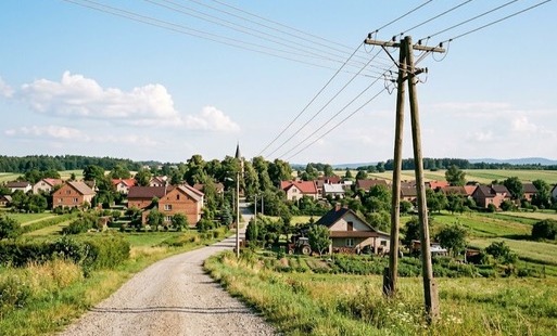 Kadr przedstawiający słup energetyczny lub linię napowietrzną niskiego napięcia na tle wiejskiego krajobrazu, w słoneczny dzień. W tle widoczne zabudowania wsi.